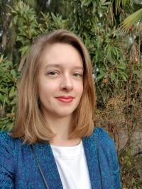 [alt text] headshot of Prof. Delmolino wearing a blue blazer with green foliage in the background
