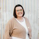 Pia Wright smiling, wearing colorful glasses and a tan cardigan.