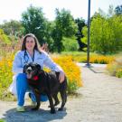 Lauren Watson, wearing a white lab coat and blue scrubs, smiles for the camera with a large black dog with its tongue hanging out.