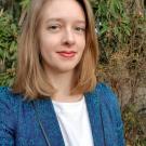 [alt text] headshot of Prof. Delmolino wearing a blue blazer with green foliage in the background